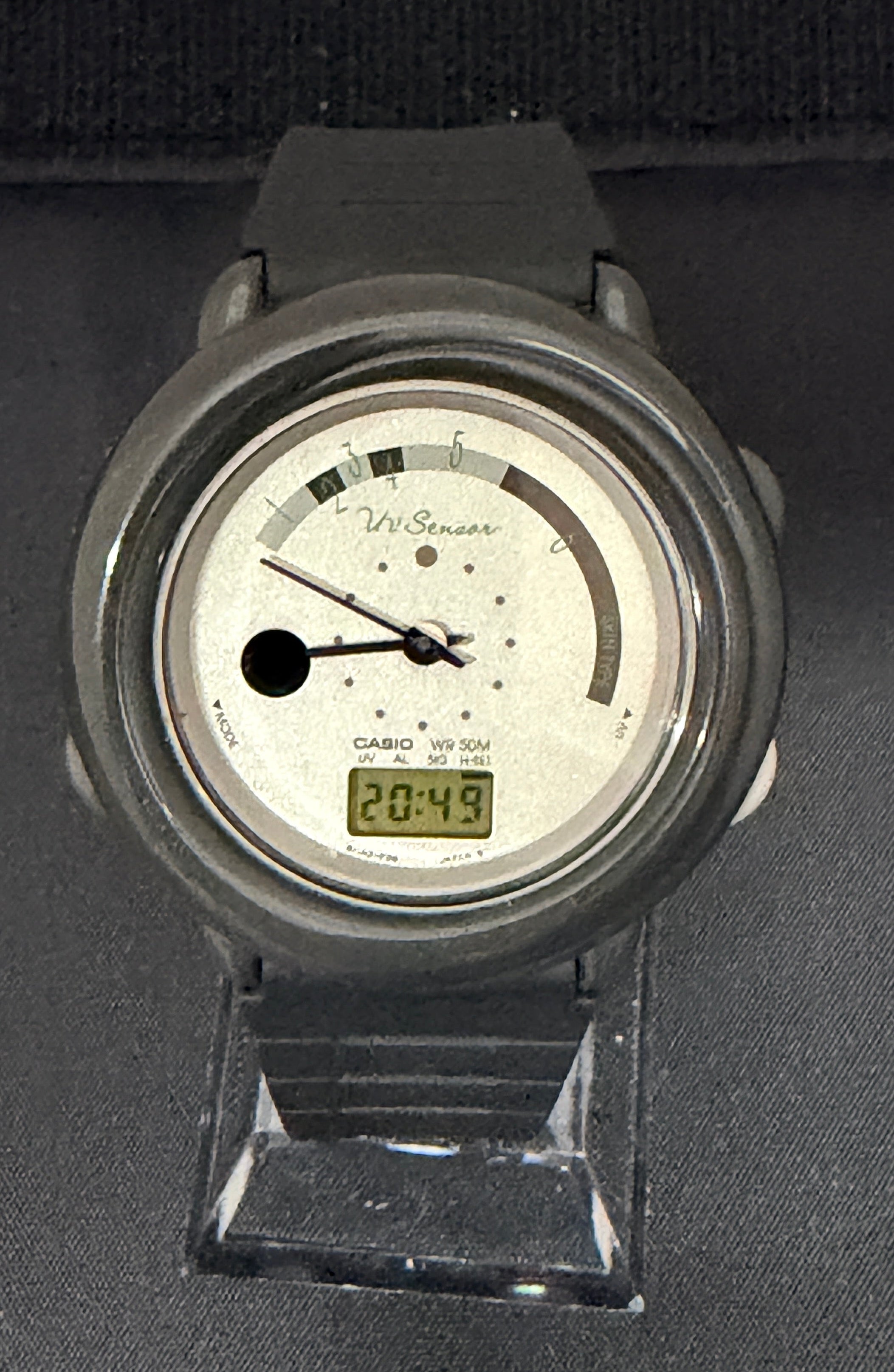 A wristwatch with a dark gray strap and case, featuring a white dial with black hands and a small green digital display, resting on a dark textured surface.