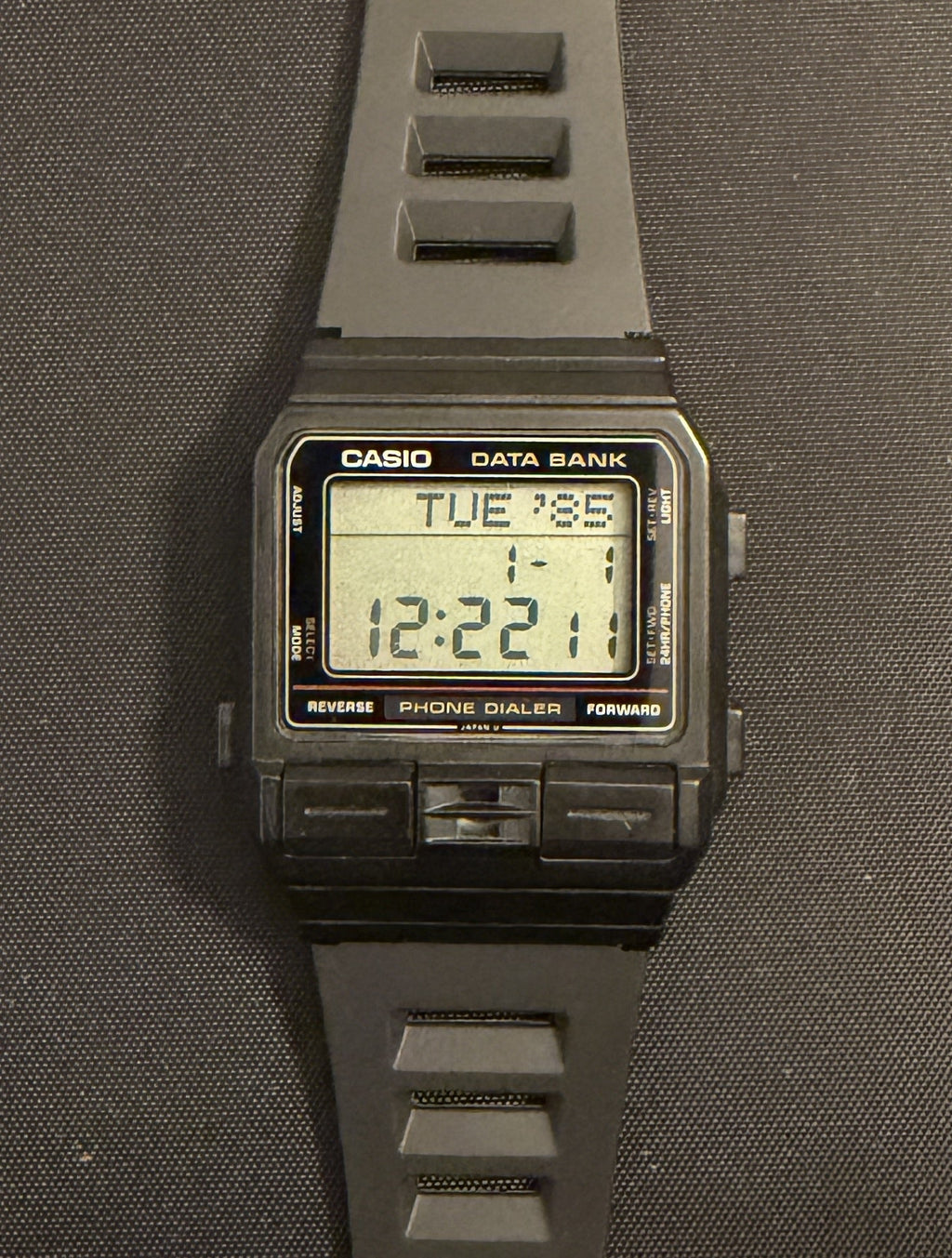 A Casio digital watch with a dark gray silicone strap and a black rectangular case, displayed vertically on a dark gray surface.