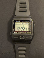 A Casio digital watch with a dark gray silicone strap and a black rectangular case, displayed vertically on a dark gray surface.