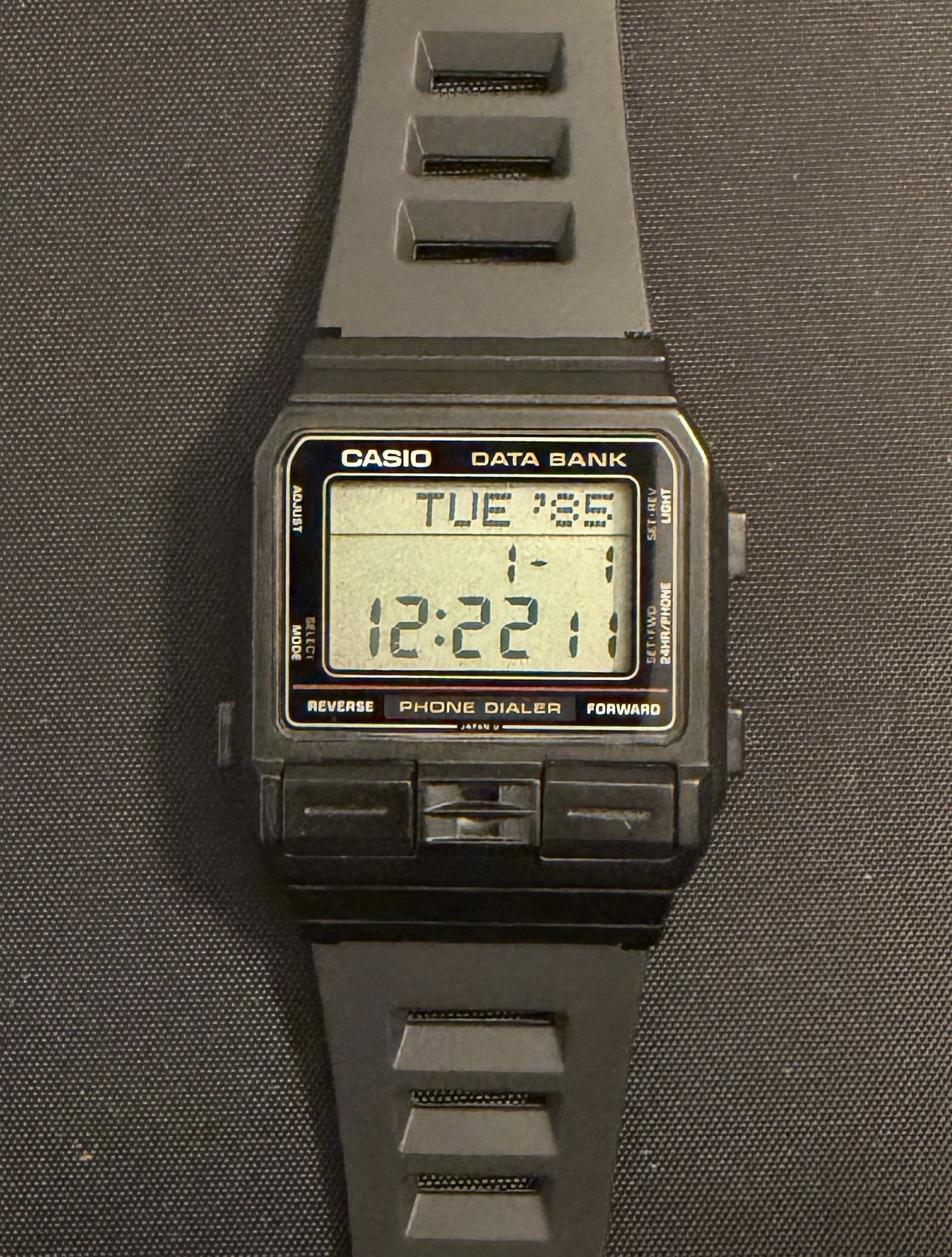 A Casio digital watch with a dark gray silicone strap and a black rectangular case, displayed vertically on a dark gray surface.