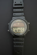 A Casio Tropicase digital watch with a black rubber strap and a black case, featuring a gray and yellow display screen, laid flat on a dark gray background.
