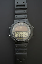 A Casio Tropicase digital watch with a black rubber strap and a black case, featuring a gray and yellow display screen, laid flat on a dark gray background.
