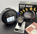 A Casio GMQ-100 Moon Graph alarm clock and matching digital watch displayed on a dark surface, with the product box and a booklet visible behind and beside them.