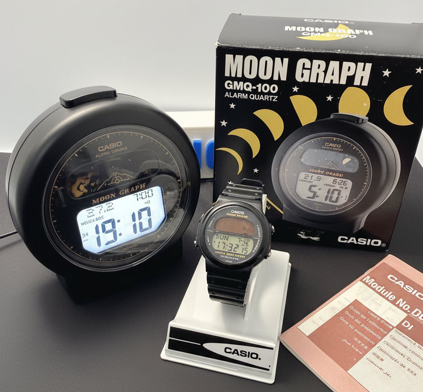 A Casio GMQ-100 Moon Graph alarm clock and matching digital watch displayed on a dark surface, with the product box and a booklet visible behind and beside them.
