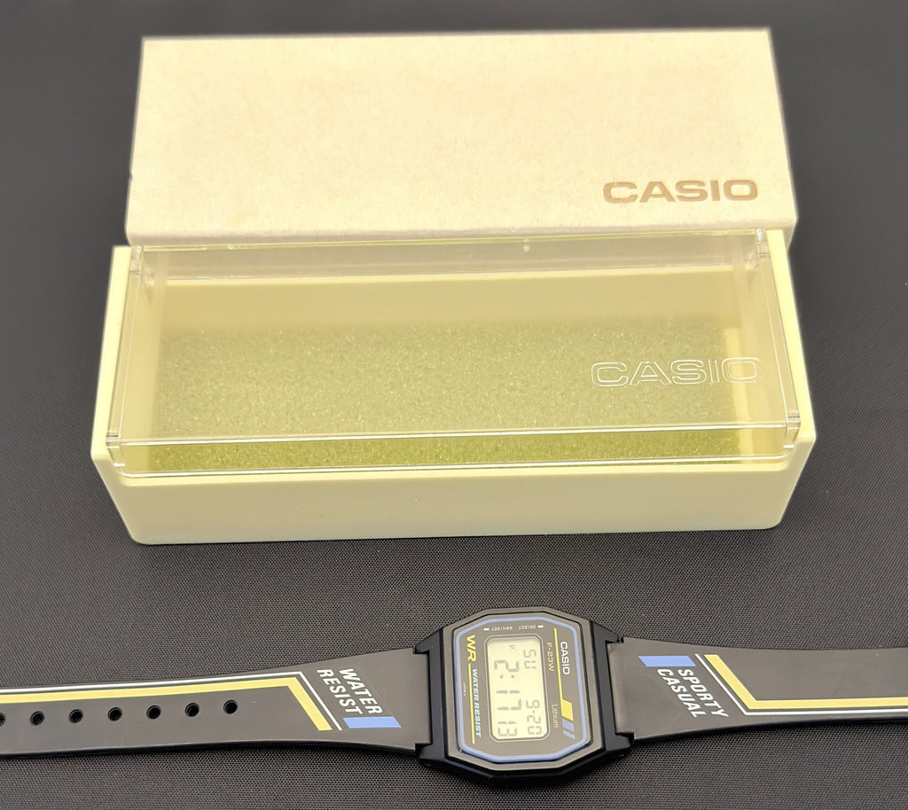 A Casio digital watch with a black strap and black rectangular case, placed horizontally below its original cream-colored Casio presentation box on a dark gray surface.