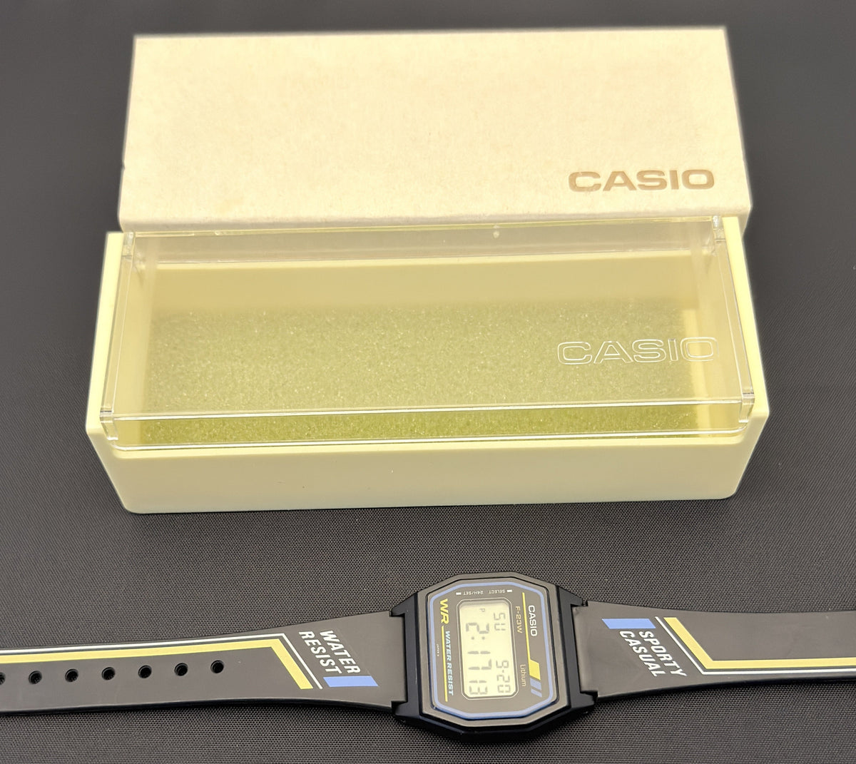 A Casio digital watch with a black strap and black rectangular case, placed horizontally below its original cream-colored Casio presentation box on a dark gray surface.