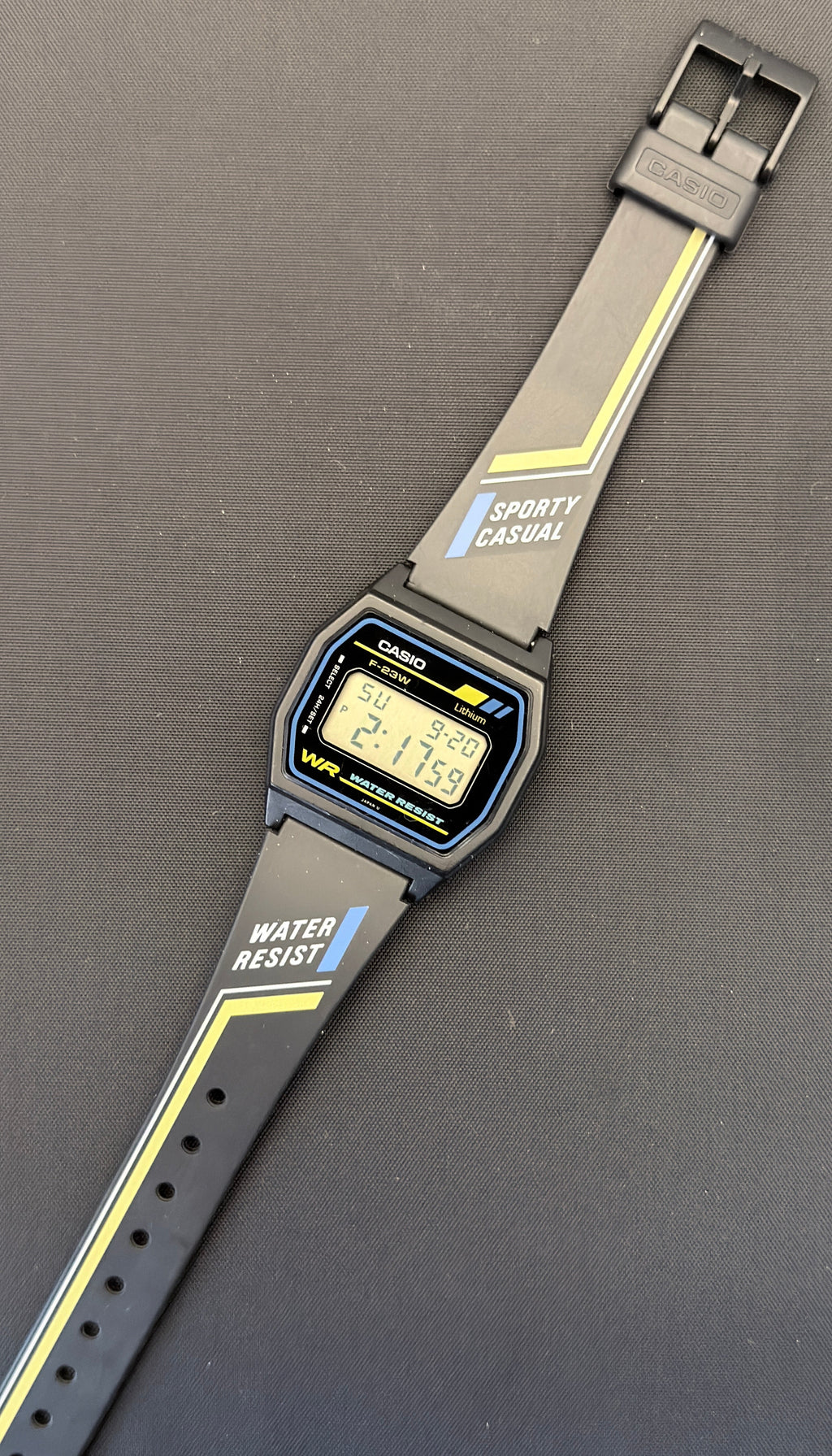 A Casio digital watch with a gray sporty strap featuring yellow stripes and white text, displayed diagonally on a dark gray surface.