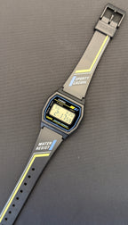 A Casio digital watch with a gray sporty strap featuring yellow stripes and white text, displayed diagonally on a dark gray surface.