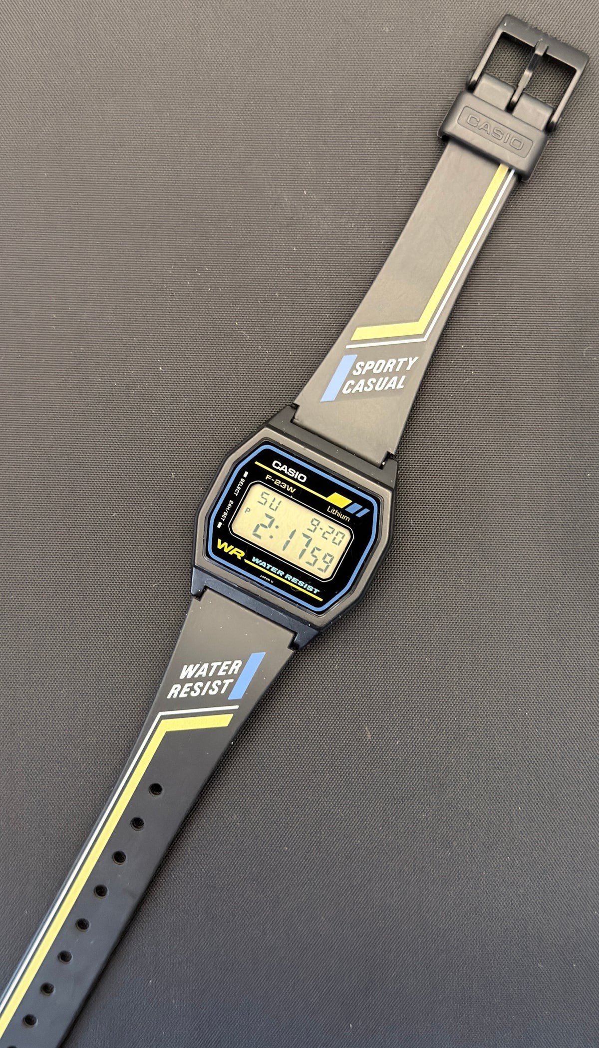 A Casio digital watch with a gray sporty strap featuring yellow stripes and white text, displayed diagonally on a dark gray surface.