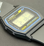 A Casio F-220W digital watch with a black case and strap, featuring a blue and yellow bezel border and a yellow 'WR' marking, resting on a textured gray surface.