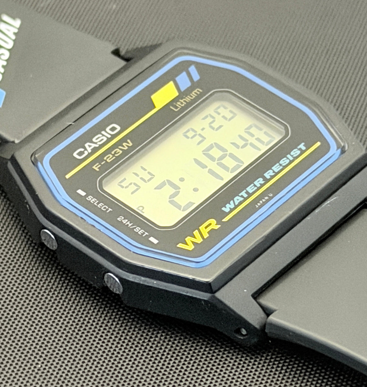 A Casio F-220W digital watch with a black case and strap, featuring a blue and yellow bezel border and a yellow 'WR' marking, resting on a textured gray surface.