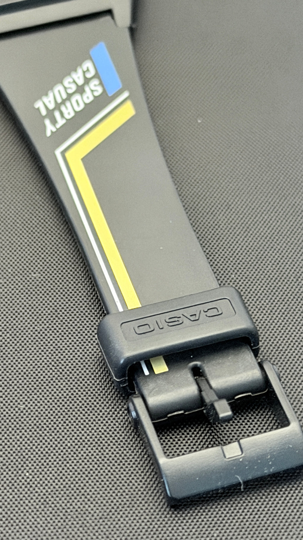 Close-up of a Casio Sporty Casual watch band in black with yellow and blue accents, featuring a black metal buckle on a textured gray background.