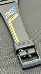 Close-up of a Casio Sporty Casual watch band in black with yellow and blue accents, featuring a black metal buckle on a textured gray background.