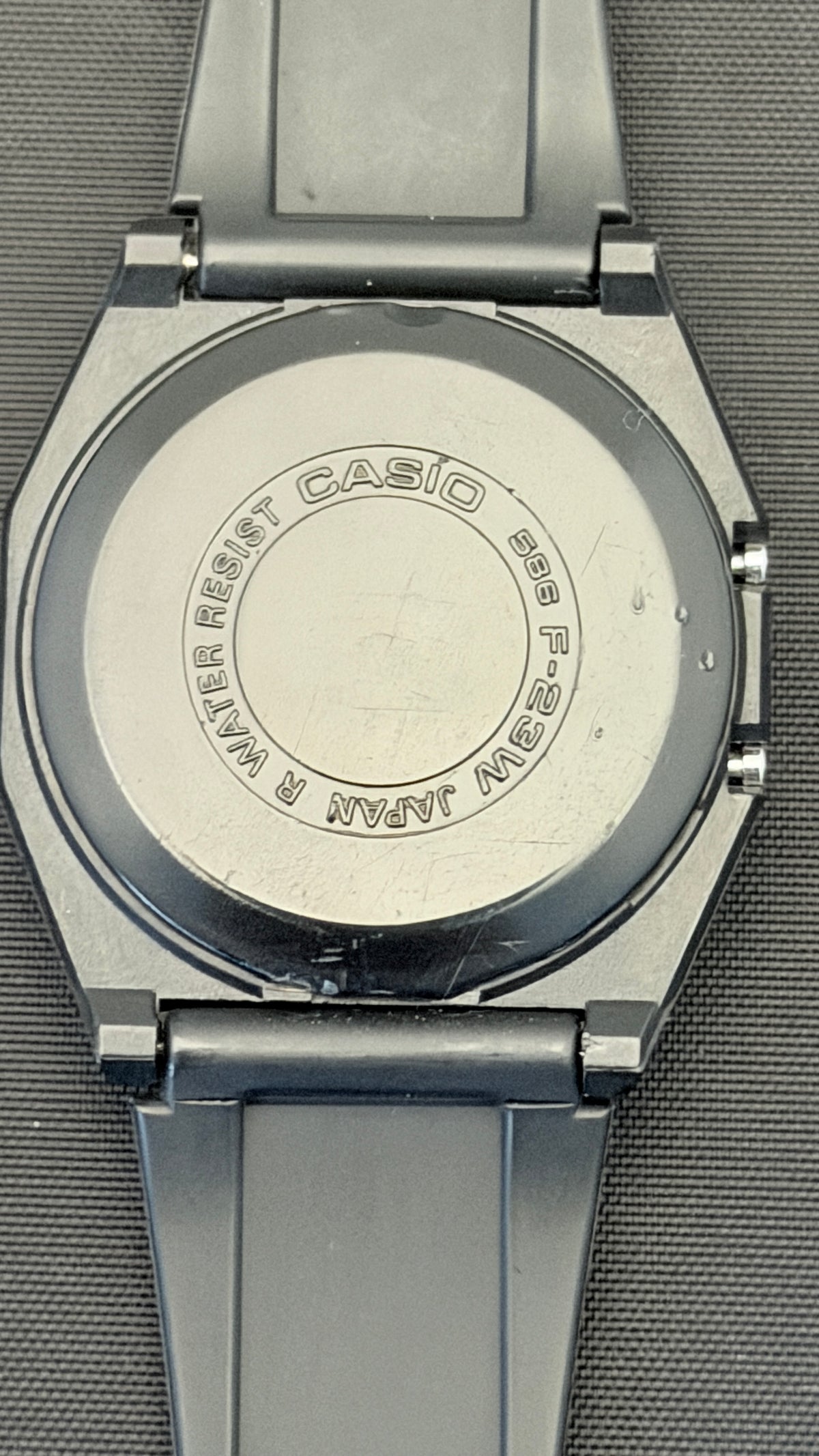 Close-up of the back of a Casio analog watch with a silver-toned metal case and dark gray strap, showing engraved text on the case back.