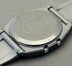 Close-up of the back of a stainless steel wristwatch with a metal link bracelet, resting on a textured gray surface.