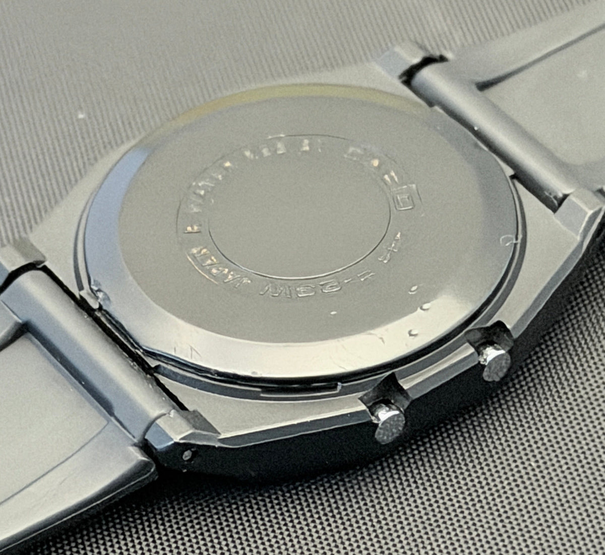 Close-up of the back of a stainless steel wristwatch with a metal link bracelet, resting on a textured gray surface.